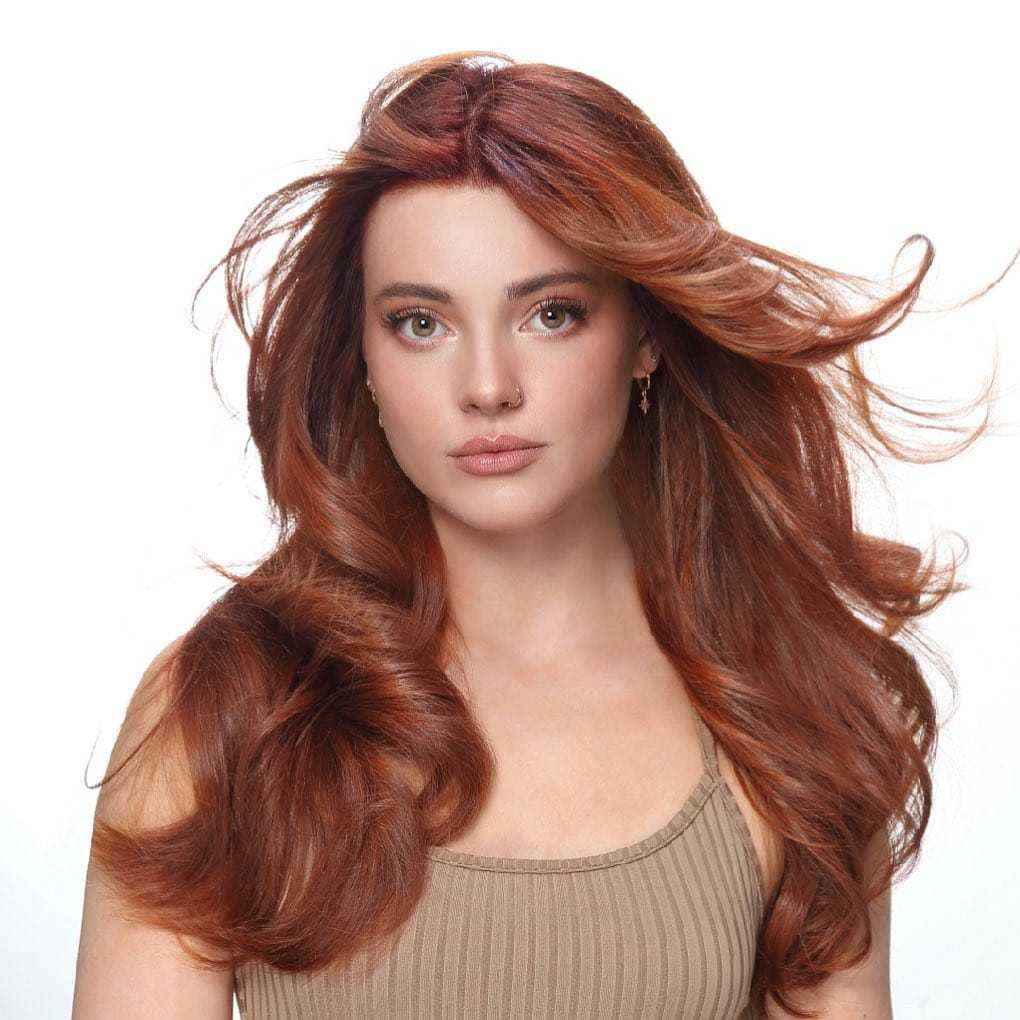 Woman with long, flowing red hair poses against white background, wearing a beige top.
