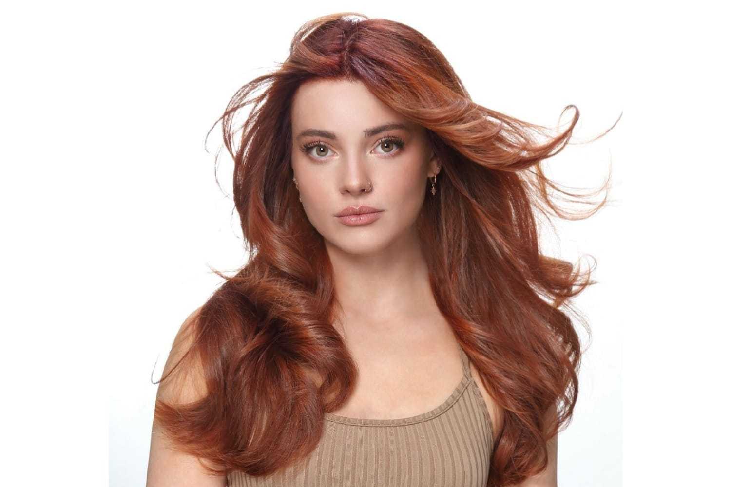 Young woman with long, flowing auburn hair, wearing a beige tank top, looking at the camera.