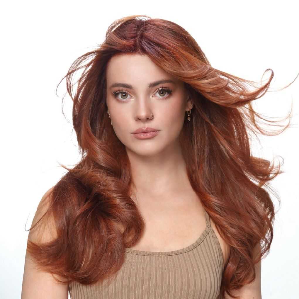 Woman with long, flowing red hair poses against white background, wearing a beige top.